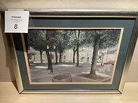 Full view of framed watercolor painting with village scene showing trees, buildings and lampposts.