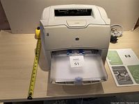 Front view of HP LaserJet 1200 printer showing paper tray with paper, and a tape measure beside for scale.
