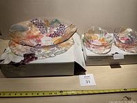 Large colored glass bowl and four smaller bowls all with fruit and floral design, displayed in branded boxes with measuring tape for scale