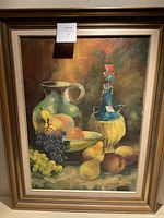 Framed oil painting showing the full scene including grapes, pears, a ceramic jug, a bowl with fruits and a wine bottle wrapped in straw. The frame is wood with gilded details and an inner cream liner.