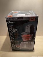 Front view of the boxed Oster food processor with product image and brand name visible.