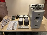 Full view of system components including five speakers, subwoofer with control unit on top, remote control, wiring, and manuals on wooden table.