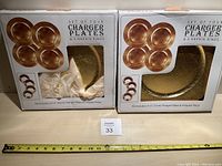 Two boxed sets of four gold charger plates each, labeled with packaging indicating size and inclusion of napkin rings, though napkin rings are missing.