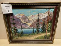 Close-up photo of a framed oil painting showing a mountain lake landscape with pine trees and a path, in a dark ornate frame.