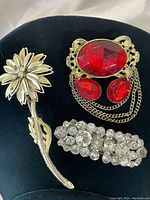 Three vintage brooches displayed together on black background: silver-tone 3D flower brooch, 1950s clear rhinestone clip, gold-tone red glass brooch with chain detail.