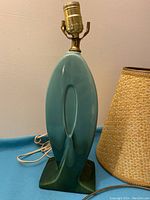 Teal oval ceramic lamp base with brass socket, partially visible woven lamp shade to right side.