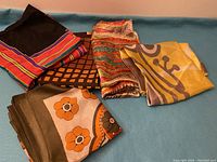 Five mixed vintage and luxury scarves arranged on a blue surface showing various patterns and colors including stripes, floral, geometric and abstract designs.