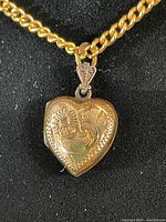 Close-up photo of heart-shaped locket showing detailed etched floral design on front, connected to bail and chain.