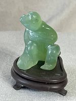 Front angled view of the translucent green jade monkey figurine sitting on a dark oval wooden stand with four small legs.
