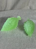 Two green vintage jadeite acorns with textured finish, displayed on a light fabric surface. The acorns vary in size and show a translucent, glossy jadeite glass look.