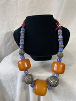 Front view of the necklace displayed on a black bust, showing the large amber beads along with smaller blue and metal beads.
