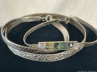 Three vintage bracelets displayed on black background showing detailed floral pattern, engraved name plate, and snake chain styles.