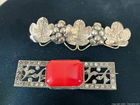 Two vintage sterling silver brooches on black background; one broad with three detailed leaves and berries, one rectangular with red cabochon stone and floral cutouts.