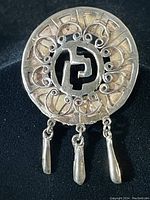 Front image showing circular pendant/brooch with geometric central design and ornamental filigree border with three dangling elements.