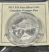 Close-up of the 2013 Canadian silver coin showing the detailed design and text: 'CANADA 10 DOLLARS 2013'. The coin depicts a summer scene by a lake with figures, a canoe, and trees.