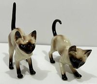 Pair of two Siamese cat ceramic figurines. One cat stands with tail straight up, the other is crouching playfully. Both have dark face, ears, paws, and tail with pale cream body and blue eyes.
