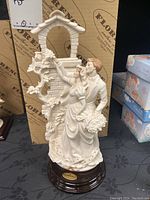 Front view of the Armani wedding couple figurine showing bride and groom near a decorative well with vines and flowers, on wooden base with nameplate.