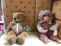 Porcelain doll and collectible teddy bear displayed side by side with original packaging in background
