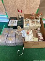 Overview of collection including Boyds figure with Canadian flag, multiple boxed and unboxed Hummel and Cherished Teddies figurines in cardboard box and on green carpet.