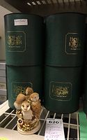 Photo of four green cylindrical original boxes stacked behind an Ivory Tower owl figurine set on a shelf.