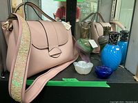 Pink leather handbag with a holographic sequin strap alongside multiple glass fragrance style lamps including blue, multicolor swirl, and cobalt blue lamps and an iridescent flower-shaped glass bowl.