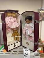 Baby doll dressed in pink gingham dress with white lace and a white shirt inside a burgundy case. Case interior contains a second pink dress and a small teddy bear.