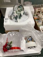 Two music boxes displayed in foam packaging: a large winter village snow globe and a smaller snowman figure music box. Also includes a decorative crystal-like candle holder with a red ribbon and flower.