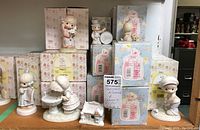 Multiple boxed and unboxed Precious Moments and Sammy's Circus figurines arranged on a shelf, showing boxed items and loose figurines including a small white ceramic house figurine.