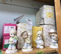 Photo showing four Precious Moments figurines with original boxes behind them including a child holding flowers, a cowboy hat child, and other characters.