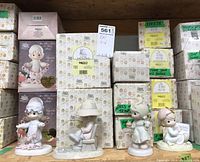 Shelf display of multiple boxed and unboxed Precious Moments figurines, including a girl with a garland of hearts, a girl sitting on a bench, and other characters in pastel colors.