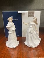 Two Royal Doulton porcelain figurines displayed with their original branded boxes behind them on a wooden surface.