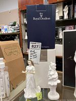 Two Royal Doulton figurines side by side with boxes in the background, one is a girl in a white dress, the other is a girl in winter attire holding a small tree