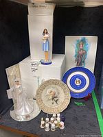 The full group of Royal Family collectibles including figurines, plates, and thimbles displayed on black fabric with some original boxes.