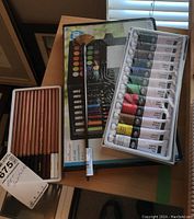 Lot contents showing Essentials oil paints, Creatology 100-piece art set, and Monte Martre colored charcoal pencils on a wooden table under window light.