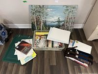 Full view of lot showing plastic tote with paints, stacked canvases, unframed landscape painting, drawing set rolled out, and loose papers/posters.