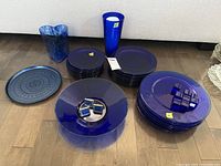 Wide shot showing stacks of cobalt blue glass plates in multiple sizes, blue patterned glass vase, tall cobalt blue glass vase, a patterned blue place card holder, cobalt blue glass bowl, metal napkin rings, and a stoneware plate.