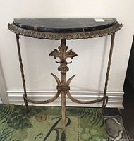 Front view showing half-moon black marble top with brownish veins and metal edging, decorative twisted metal legs, and fleur-de-lis motif on base.