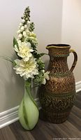 Full view of two floor vases side by side on floor; green swirl glass vase with faux white flowers, and large brown ceramic jug vase with intricate designs.