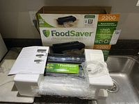 Photo showing FoodSaver vacuum sealing system, original box, manual, vacuum hose, and protective styrofoam packaging on sink counter.