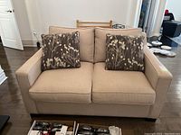 Front view of beige upholstered love seat with two patterned throw pillows on hardwood floor