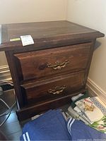 Two images showing front and angled views of a wood veneer night table featuring two drawers with decorative metal pulls, placed near a corner wall with folded linens beside it.
