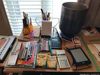 View of the lot showing Kobo eReader with cover, assorted stationery items, two calculators, crayons, stapler in packaging, pens in holders, spiral-bound address book, ruler, metal bin and other miscellaneous items.