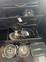 Open kitchen drawer showing assorted metal pans, pots, and lids, some items placed on floor in front.