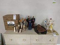 Full view of religious figures displayed on white dresser, including Jim Shore Black & Gold nativity set with original box, wood-carved nativity piece, other religious statues and wall art pieces.