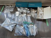 Wide layout of all flatware pieces including forks, spoons, knives, and serving utensils arranged on floor, many sealed in plastic bags with two original blue Oneida boxes visible.