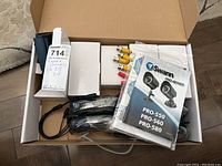 Open box showing two black Swann security cameras, cables and connectors, user manual, and packaging.