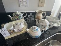 Overview of all items on a black granite countertop near a sink. Multiple tea pots with floral designs, a gravy boat, creamware jug, and teapot with cup set visible.