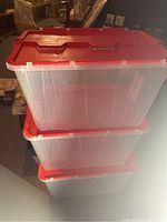 Stack of three transparent plastic storage totes with red flip-top lids, viewed from the side showing the handles and locking clips on the lids.