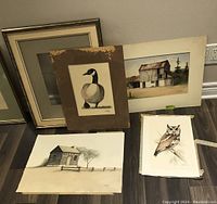 Lot showing multiple unframed signed paintings leaning against wall: barn, owl, goose, cabin and one empty frame. Visible wear and damage to mattes.