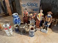 Overall view of 9 barware and collectible items arranged on floor with backdrop of cardboard box and wooden beams.